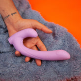 A hand holds the lavender Cute Little Fuckers Jix, a U-shaped silicone toy with an ergonomic handle, against a fluffy blue fabric backdrop. The scene rests on a red surface, and the person's wrist features a tattoo—offering an inviting introduction to this beginner anal toy by Cute Little Fuckers.